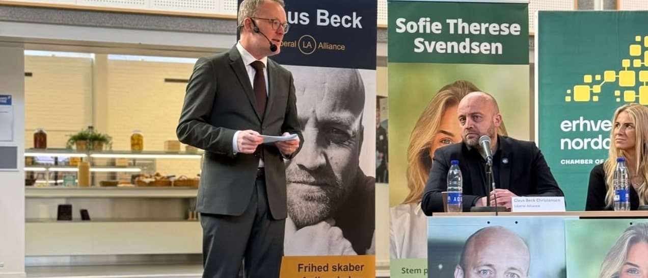 Claus Beck i panelet ved et debatmøde, med sin roll-up i baggrunden